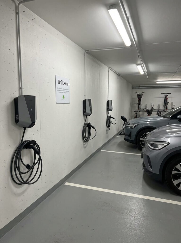 Parkeringsplats inomhus med Laddbox laddningsstationer för elfordon monterade på en vit vägg. Två gråa bilar är anslutna till laddarna. På väggen sitter en skylt med texten "Brf Eken" ovanför en grön logotyp och i taket lyser lampor upp utrymmet.